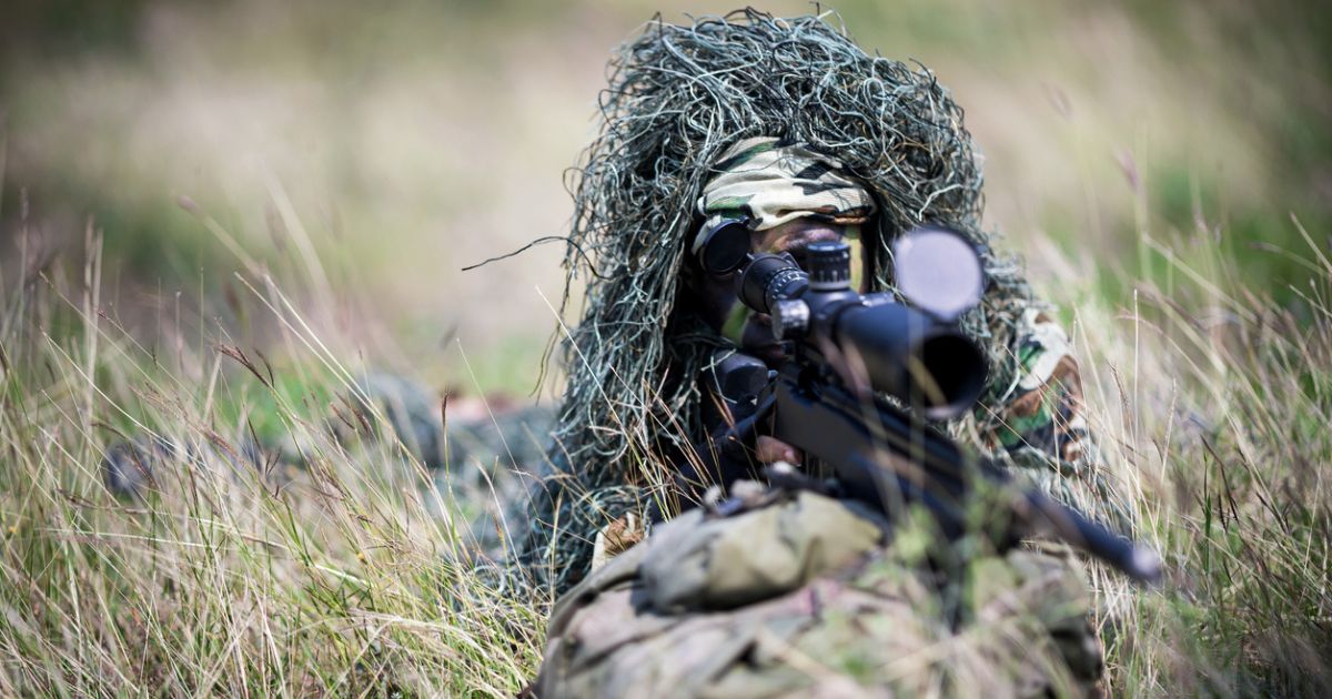A sniper hiding in brush using different signature reduction techniques to hide herself.