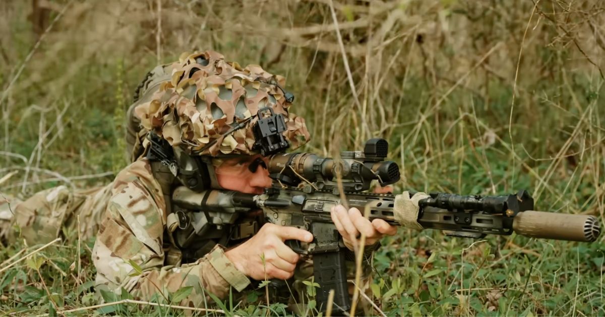 A man laying in the grass covered in camo lining up his shot with his rifle.