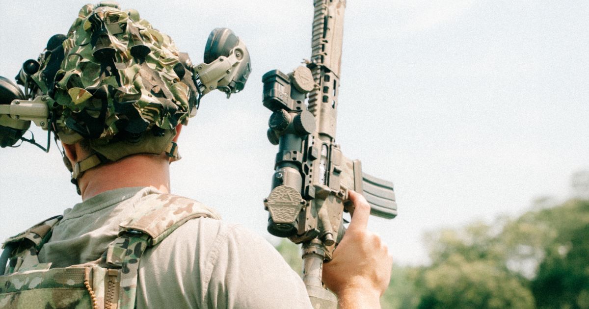 A man holding his rifle while wearing a helmet, scrim, and ear protection.
