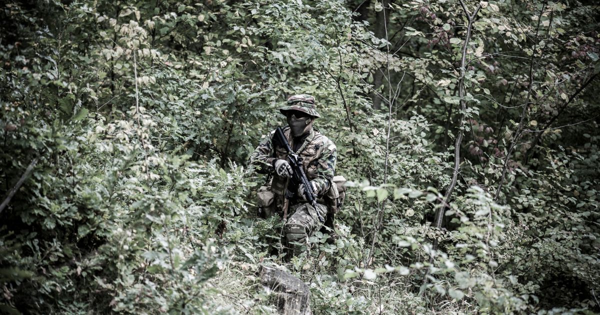 A person wearing camo holding a rifle standing in bushes.