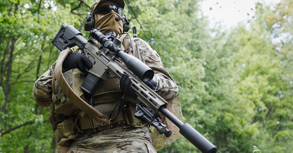 A long distance Army shooter standing in full camo with his rifle and gear pouches.