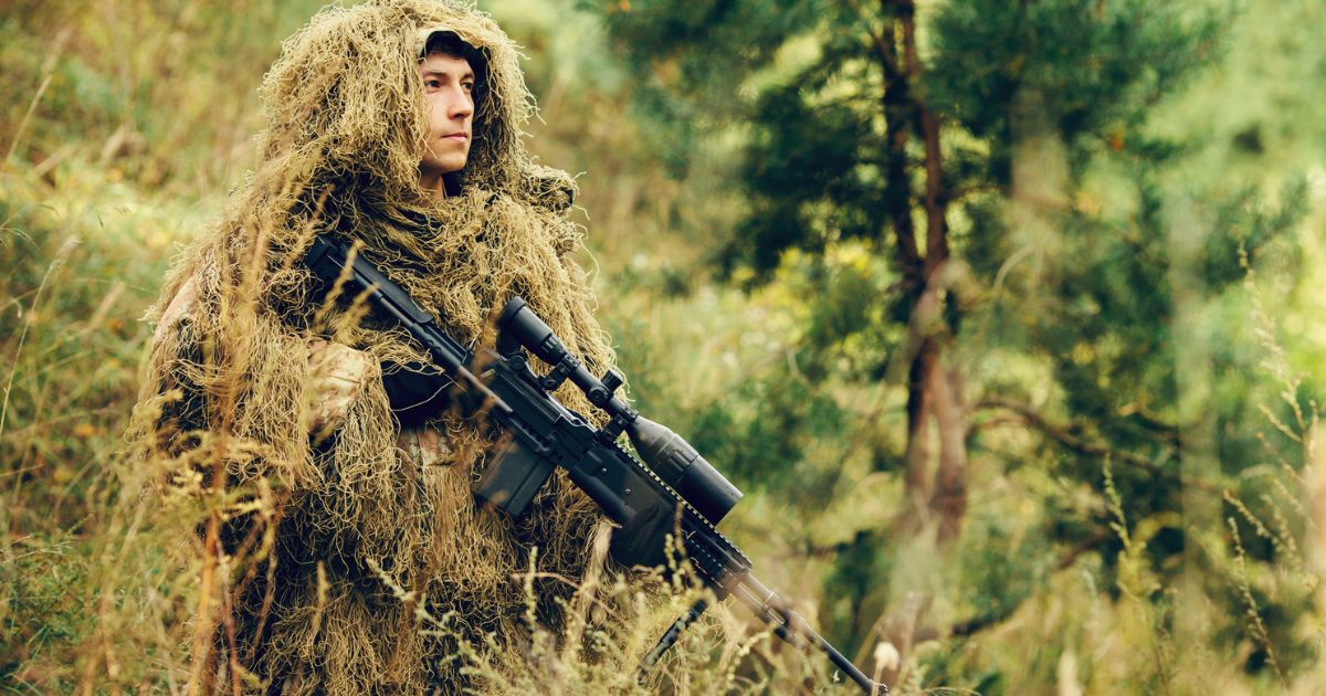 A sniper standing with his rifle in a ghillie suit.