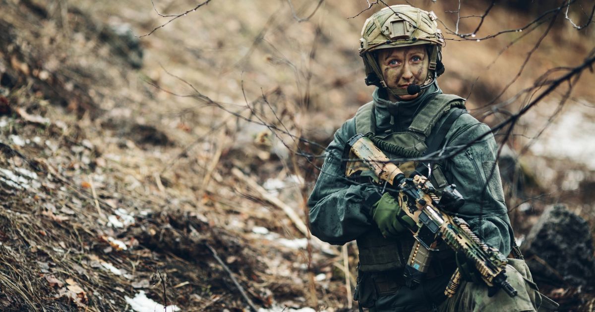 A woman sniper walking through a snowy, rocky terrain.